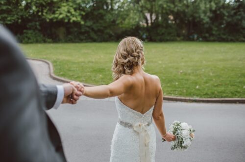 bride in backless wedding gown holding bouquet while holding grooms hand from behind